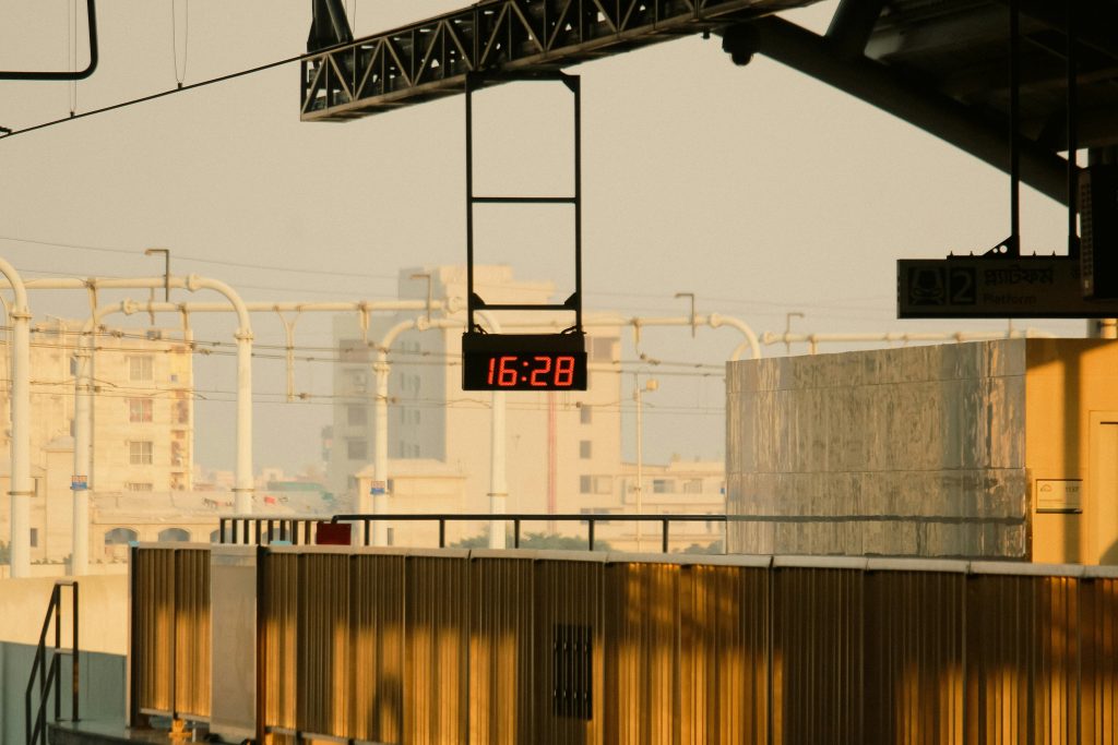 Digital clock displaying 16:28 at a railway station during golden hour, illuminated by warm sunset light. Horloge numérique affichant 16:28 dans une gare ferroviaire à l'heure dorée, illuminée par la lumière chaude du coucher de soleil.