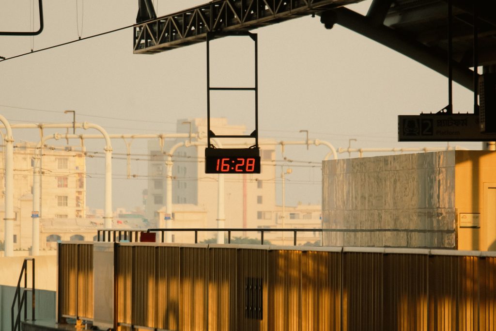 Digital clock displaying 16:28 at a railway station during golden hour, illuminated by warm sunset light. Horloge numérique affichant 16:28 dans une gare pendant l'heure dorée, illuminée par la lumière chaude du coucher de soleil.