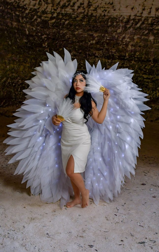 A woman in a white angelic costume with large feathered wings poses indoors. Perfect for fantasy themes. Une femme en costume angélique blanc avec de grandes ailes à plumes pose en intérieur. Parfait pour les thèmes fantastiques.