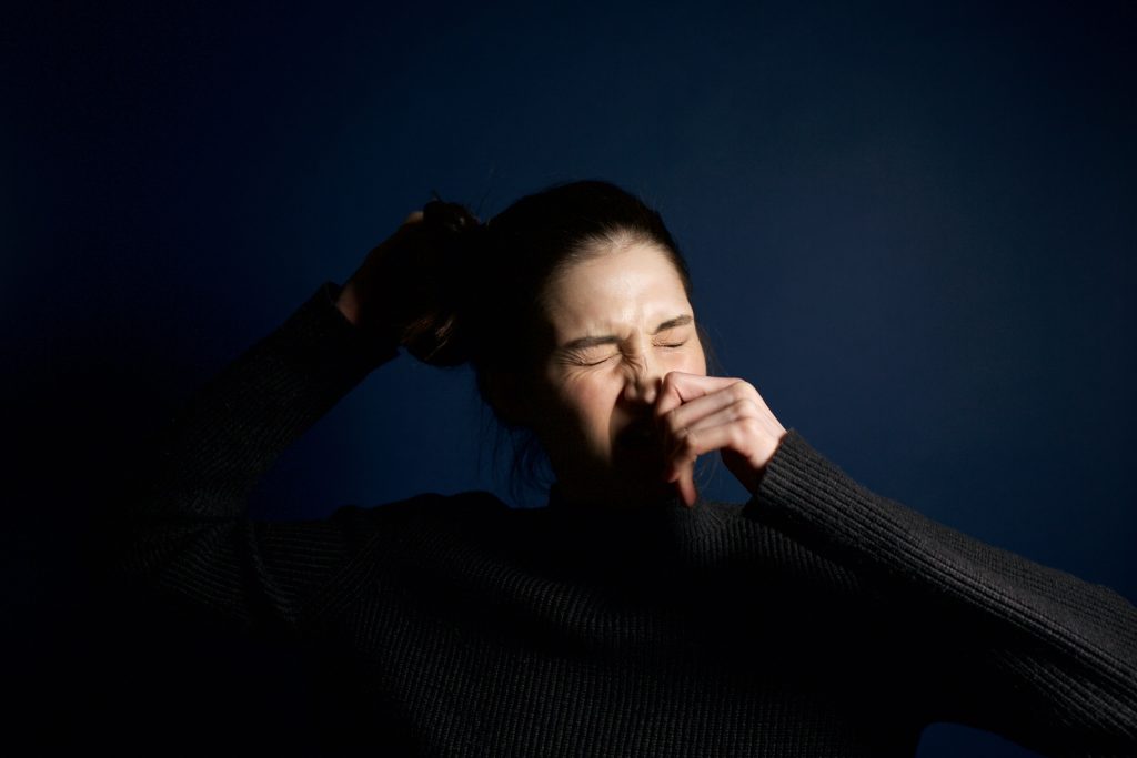 A woman in a dark sweater sneezes against a solid blue background, capturing a moment of candid expression. Une femme en pull foncé éternue sur un fond bleu uni, capturant un moment d'expression spontanée.