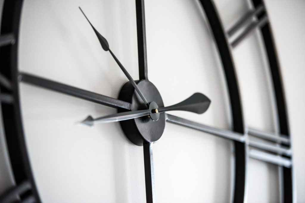 Stylish black round shaped clock with hands and Roman numeral hanging on white wall in light room on blurred background Une horloge murale ronde élégante de forme noire avec des aiguilles et des chiffres romains accrochée à un mur blanc dans une pièce lumineuse sur fond flou.