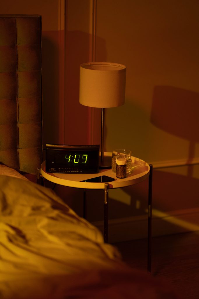 Warmly lit nightstand with lamp and digital alarm clock at 4:07 AM. Une table de nuit chaleureusement éclairée avec une lampe et un réveil numérique affichant 4:07 du matin.