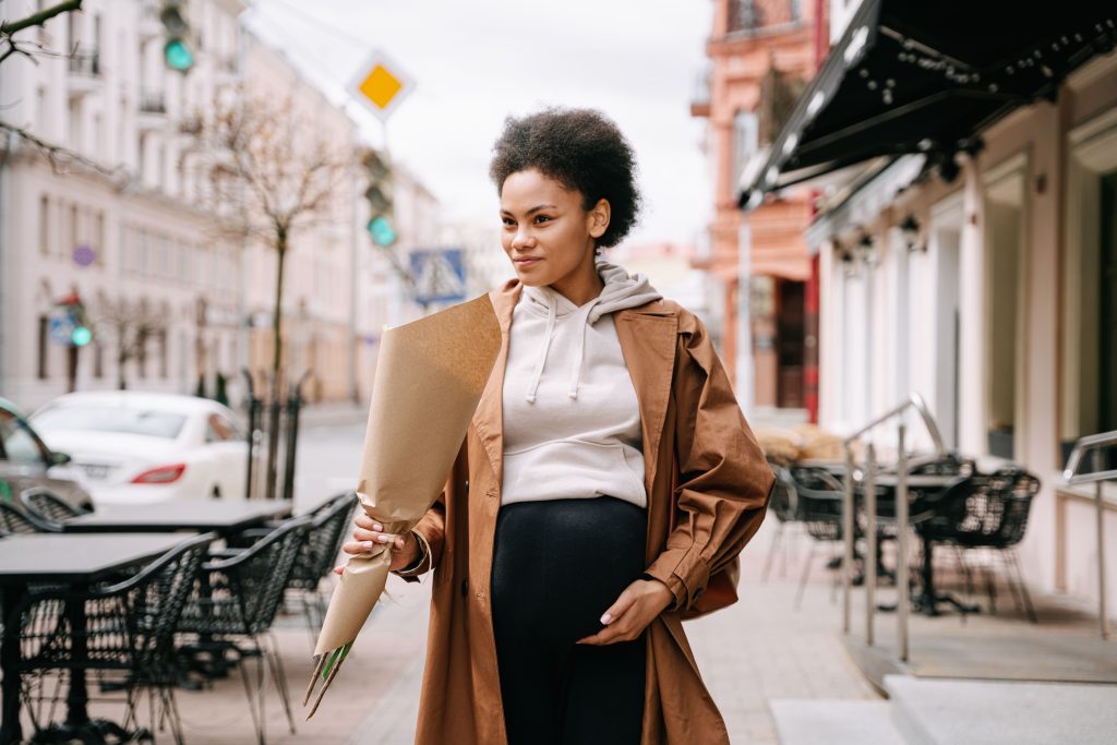 A stylish pregnant woman with afro hair holding flowers, walking down a charming city street. Une femme enceinte élégante aux cheveux afro tenant des fleurs, marchant dans une charmante rue de la ville.